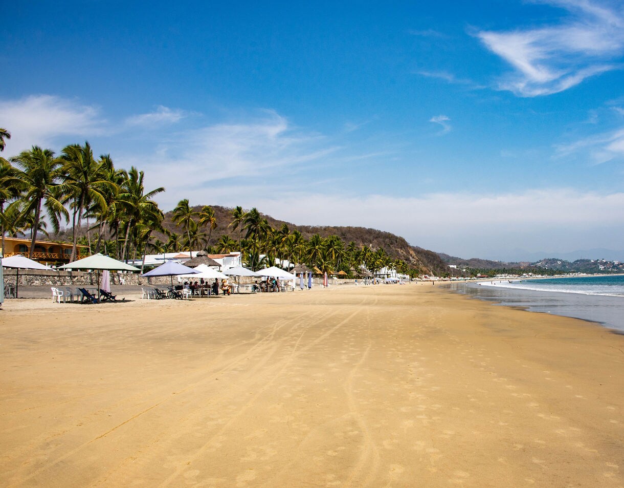Wide sandy beach in Playa Santiago, Manzanillo, lined with palm trees, white umbrellas and people wading in the calm ocean.