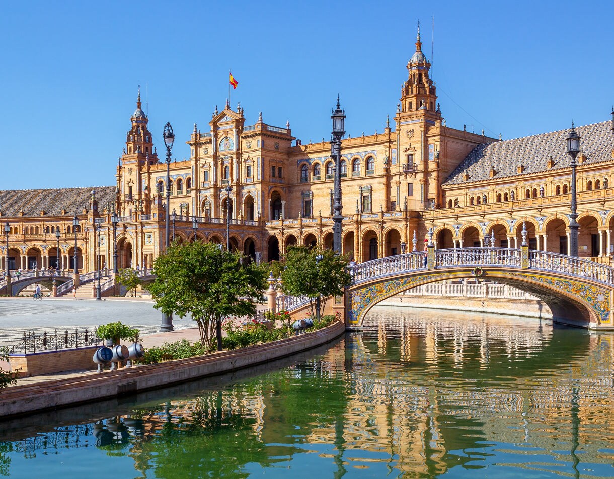 Wide view of Plaza de España in Seville showing the grand curved palace, arched colonnades and a decorative ceramic bridge reflecting in the green canal water under a clear blue sky.