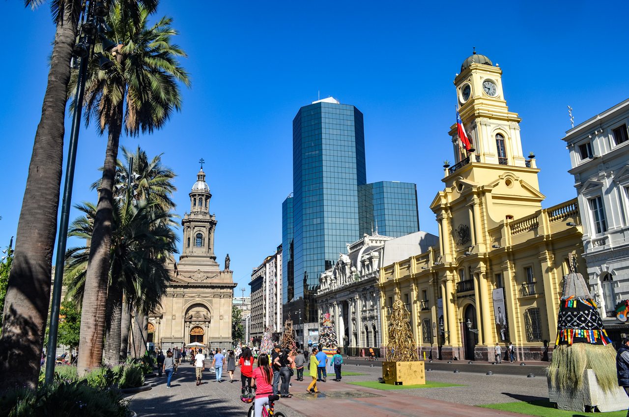 People walk through Plaza de Armas in Santiago surrounded by palm trees, a historic cathedral, a yellow clock tower and modern glass skyscrapers.