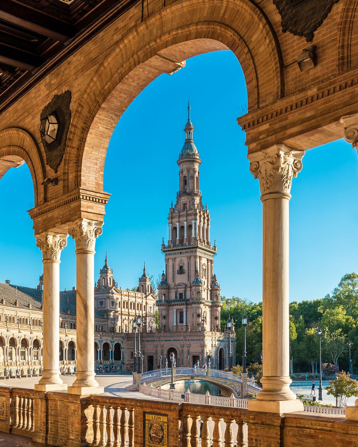 View of a tall tower at Plaza de España in Seville seen through brick arches and marble columns, with a decorative bridge and reflecting water below under a clear blue sky.
