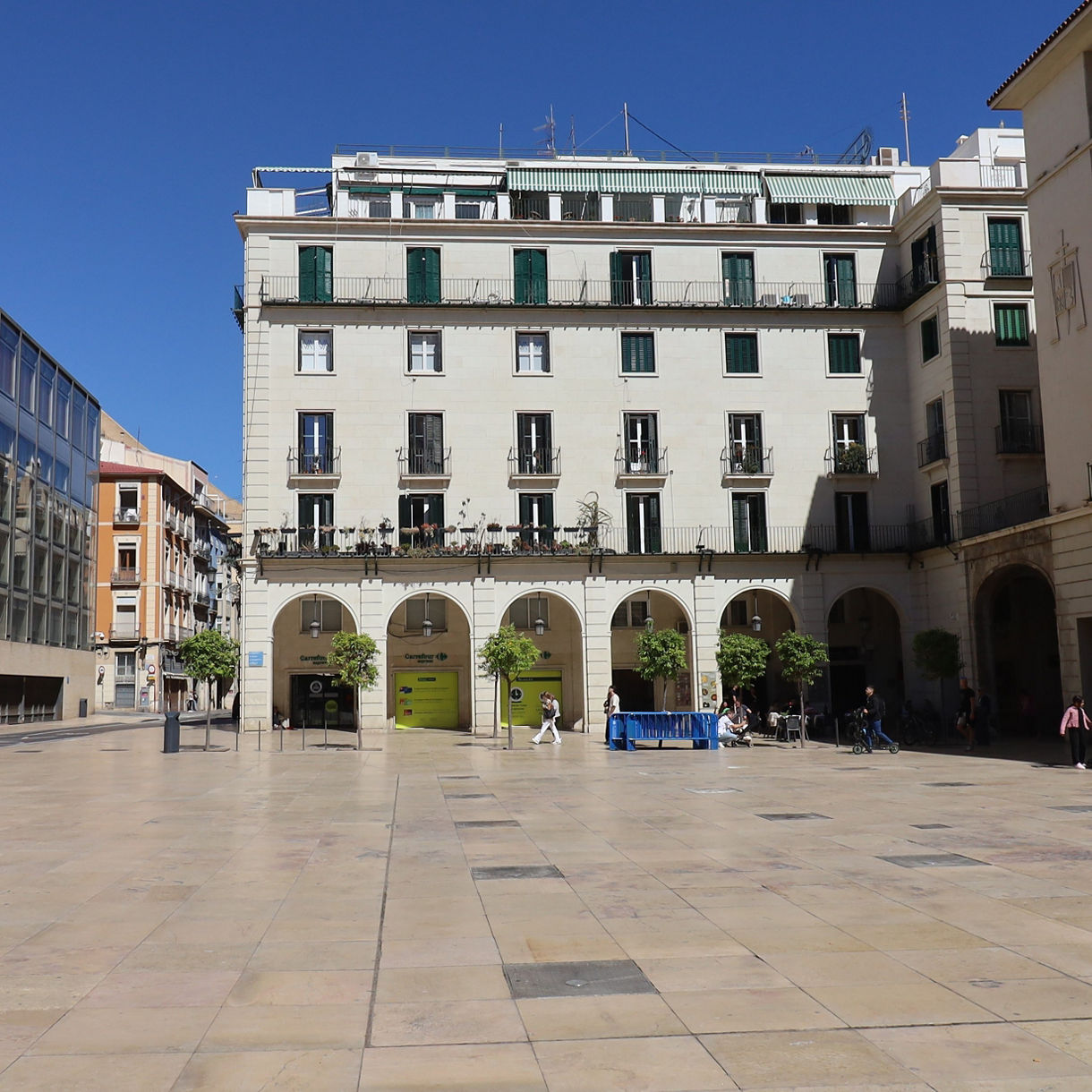 Wide plaza in Alicante lined with pale stone buildings, arched walkways and green-shuttered windows, with people walking across the open square under a clear blue sky.