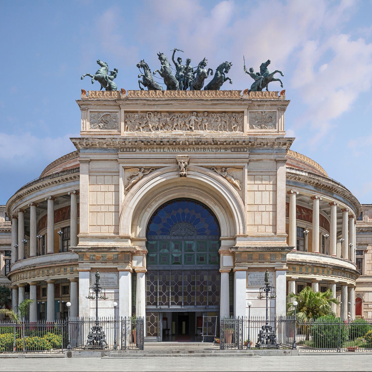 Front view of the Politeama Garibaldi theater in Palermo, showing its large arched entrance, columned façade and bronze chariot sculpture on the rooftop.