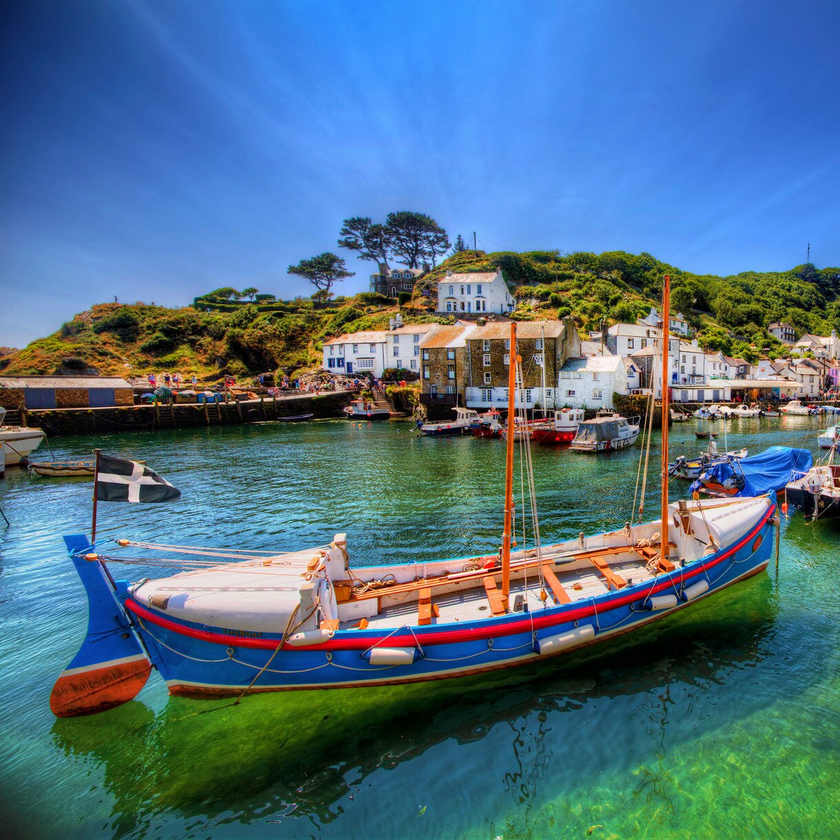 Bright fishing boats floating in Polperro’s small harbor, with whitewashed cottages and green hills rising behind under a vivid blue sky.