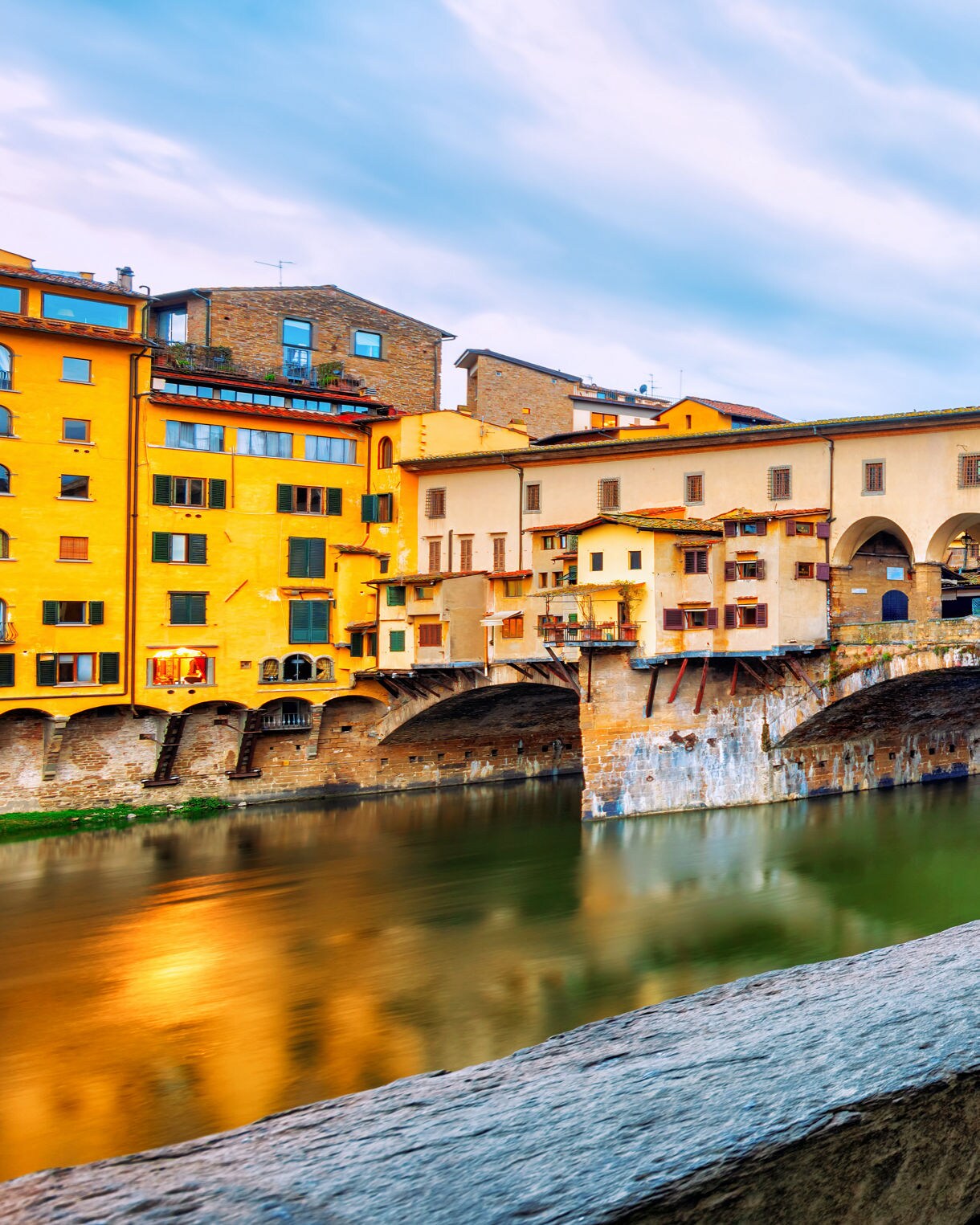 The Ponte Vecchio bridge in Florence with colorful buildings built along it, viewed from a covered stone walkway lined with repeating arches beside the river.