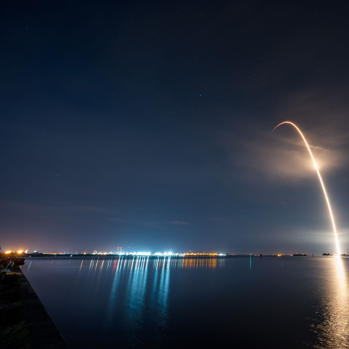 A bright rocket streaks upward into the night sky, leaving a curved trail of light reflected on the still water below. City or launch site lights glow along the distant shoreline, adding colorful reflections to the scene. Wispy clouds partially veil the sky, enhancing the dramatic arc of the rocket’s ascent. The overall atmosphere is serene yet dynamic, combining peaceful waters with the energy of spaceflight.