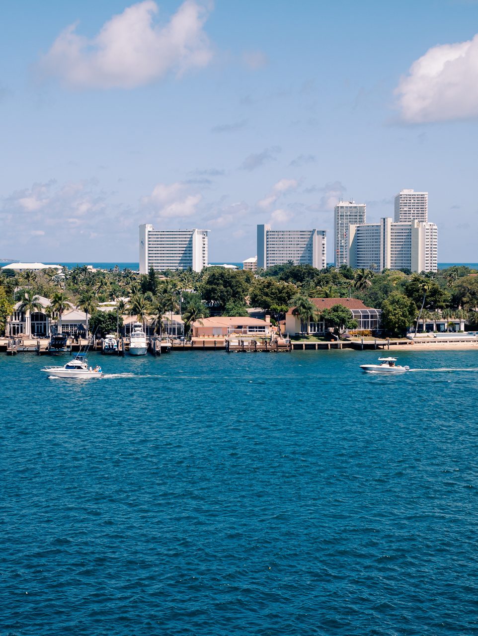 Boats cruising near waterfront residences in Port Everglades with tall hotel buildings and palm trees in the background.