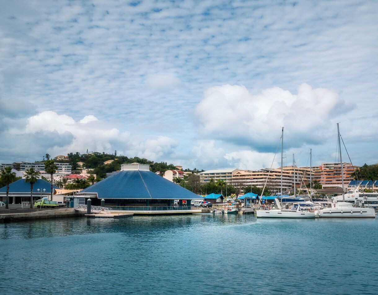 Marina scene at Port Moselle with sailboats docked beside blue-roofed market buildings, backed by low hills and pastel apartment blocks under a partly cloudy sky.