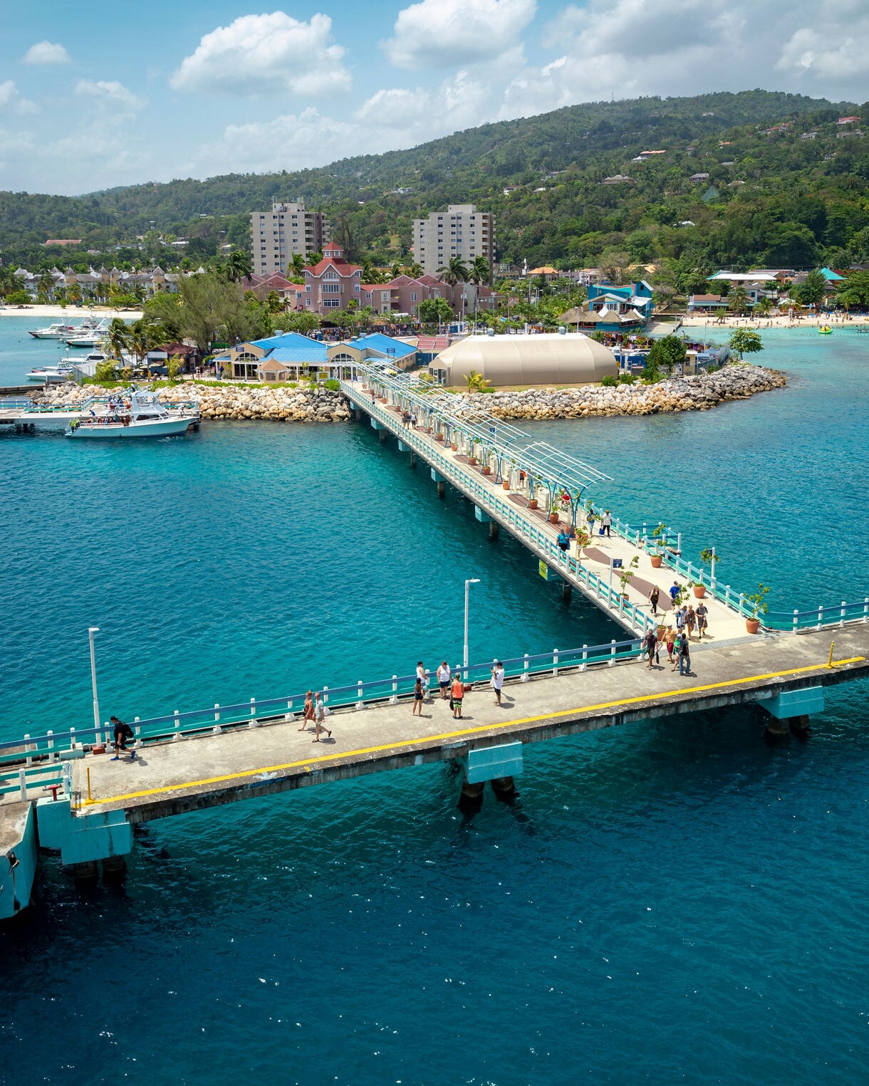 A pier extending into turquoise waters with tourists walking toward the colorful waterfront buildings and green hills of Ocho Rios.