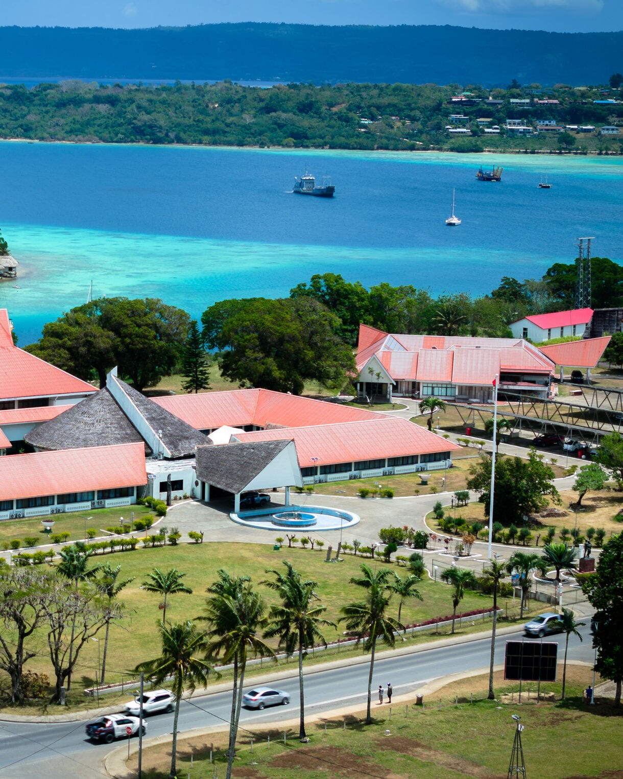 Aerial view of Port Vila’s harbor with red-roofed buildings, palm-lined roads and turquoise water surrounding nearby islands.