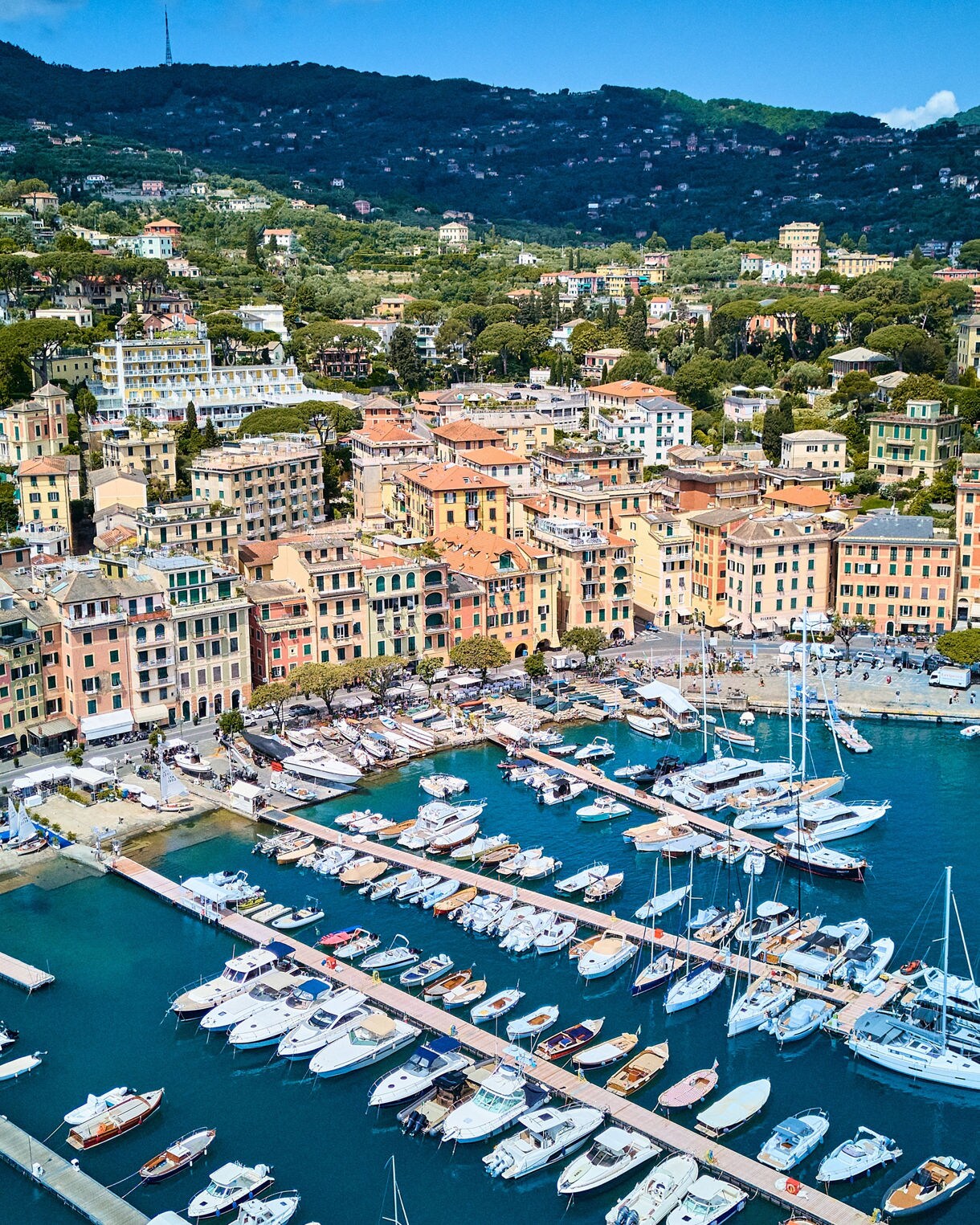 Aerial view of a colorful seaside town with a busy marina filled with boats and green hills rising behind.