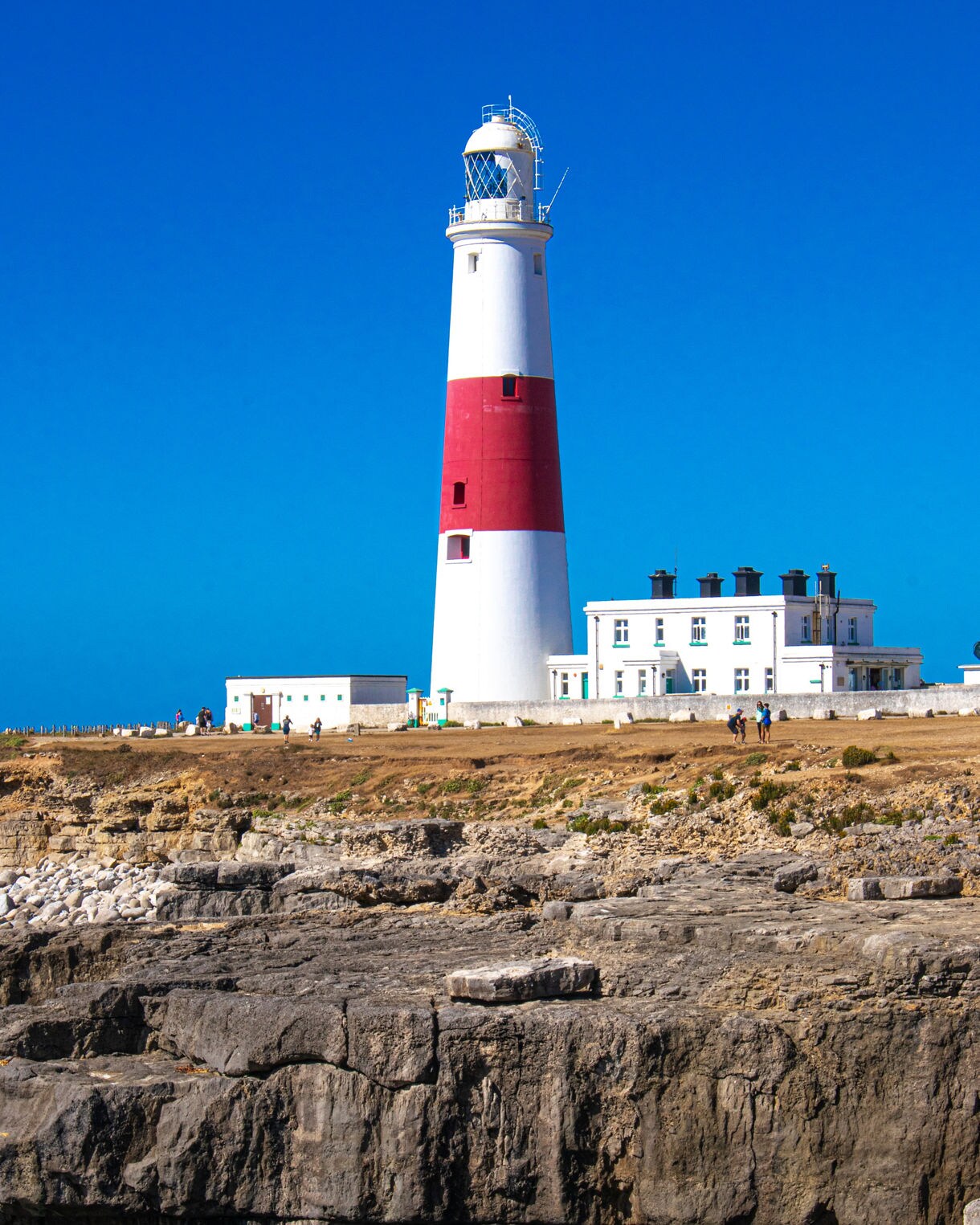 Portland Bill Lighthouse in Dorset, a tall white tower with a red central band, standing on rocky cliffs against a bright blue sky.