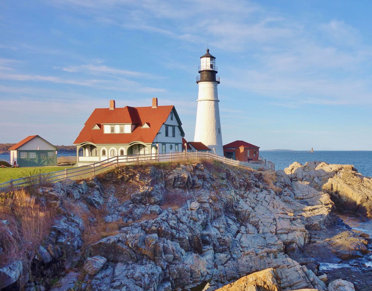 Portland Head Light perched on rocky cliffs overlooking a calm blue ocean, with a red-roofed keeper’s house beside it under a clear sky.