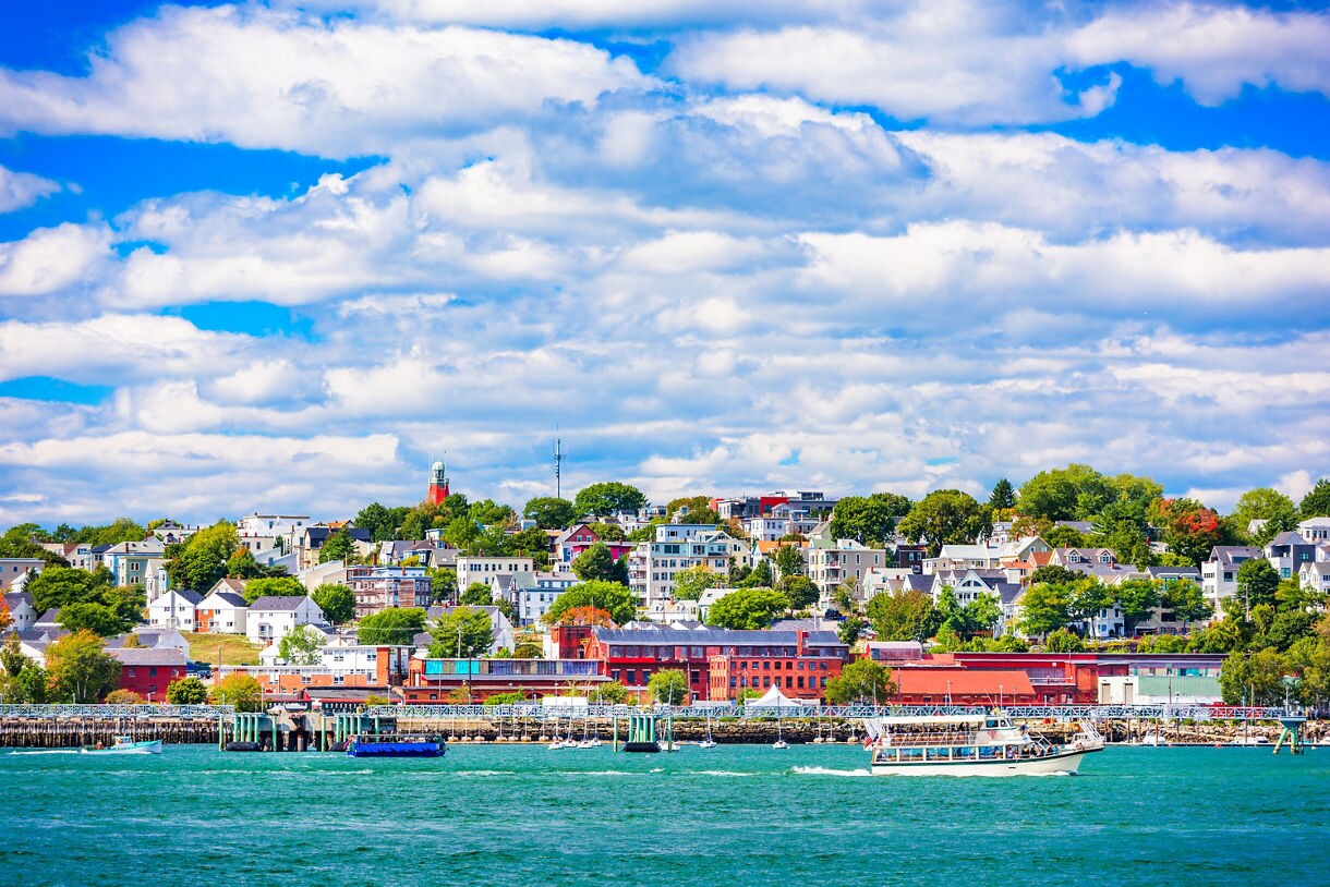 A view of Portland’s waterfront with boats moving across teal water and rows of colorful houses rising on a green hillside below a sky filled with soft white clouds.