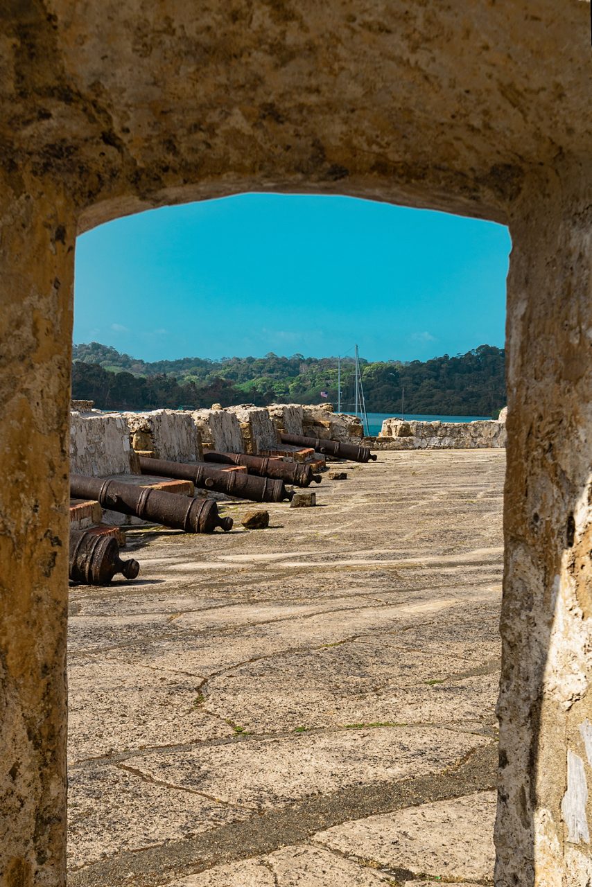 Old cannons lined along the stone fortifications of Portobelo ruins in Panama, with forested hills and sea in the background.