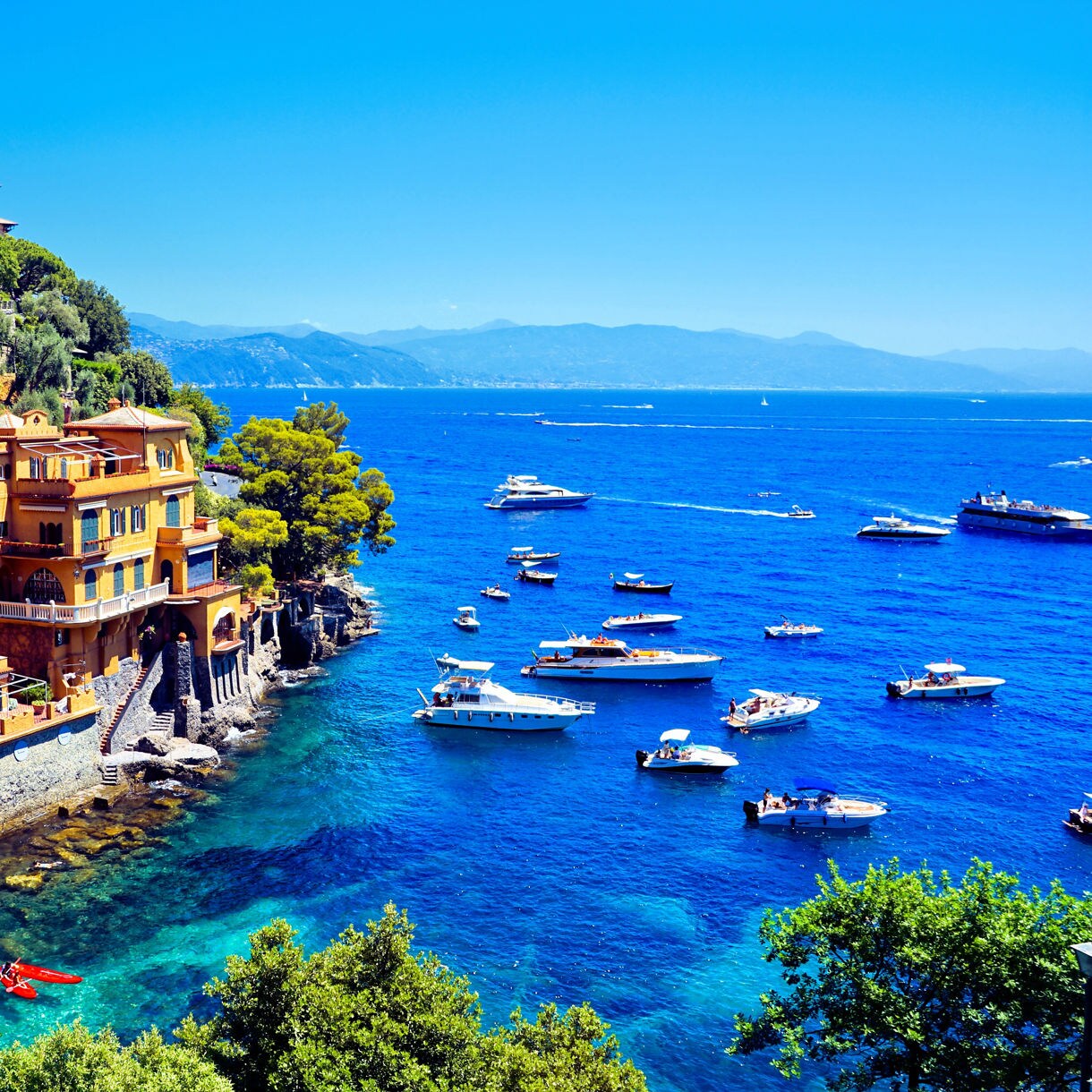 Colorful villas perched on a rocky coastline above bright turquoise water, with numerous small boats floating offshore near lush green hills.