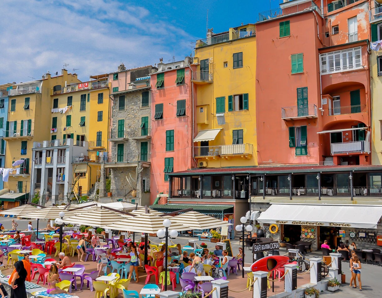 Rows of tall, colorful buildings with green shutters line a lively waterfront, where people relax at outdoor café tables under striped umbrellas, near the rocky shore.