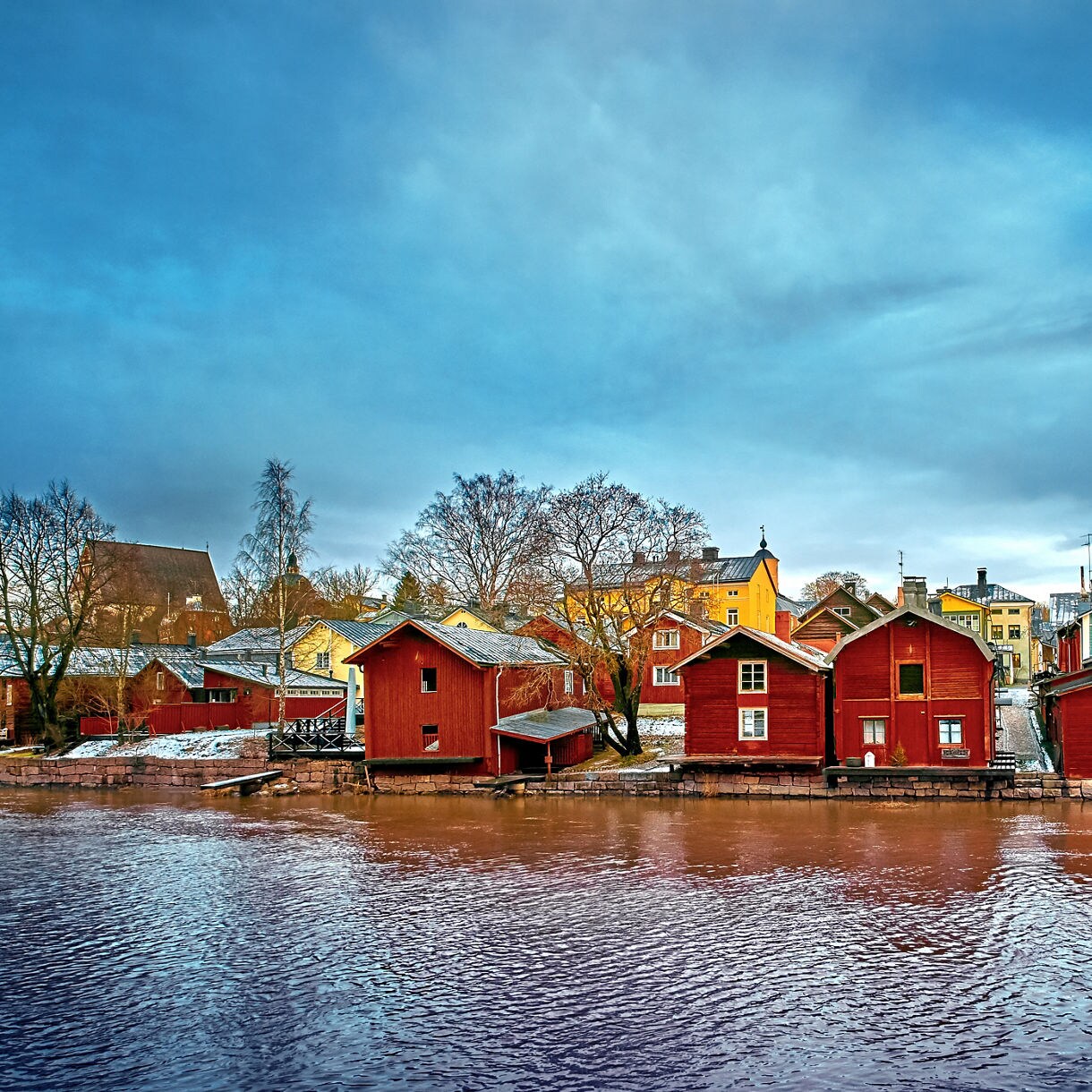 Traditional red wooden riverside houses in Porvoo, Finland, with colorful village buildings and snow-dusted rooftops under a cloudy winter sky.