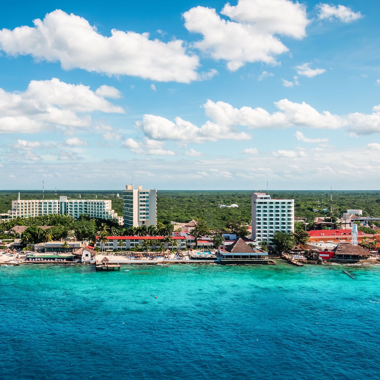 An elevated wide shot of the Cozumel coastline featuring bright blue Caribbean waters, white sandy beaches, several multi-story hotels, and a large thatched pyramid-style building nestled against a vast green forest under a bright sky with scattered white clouds.