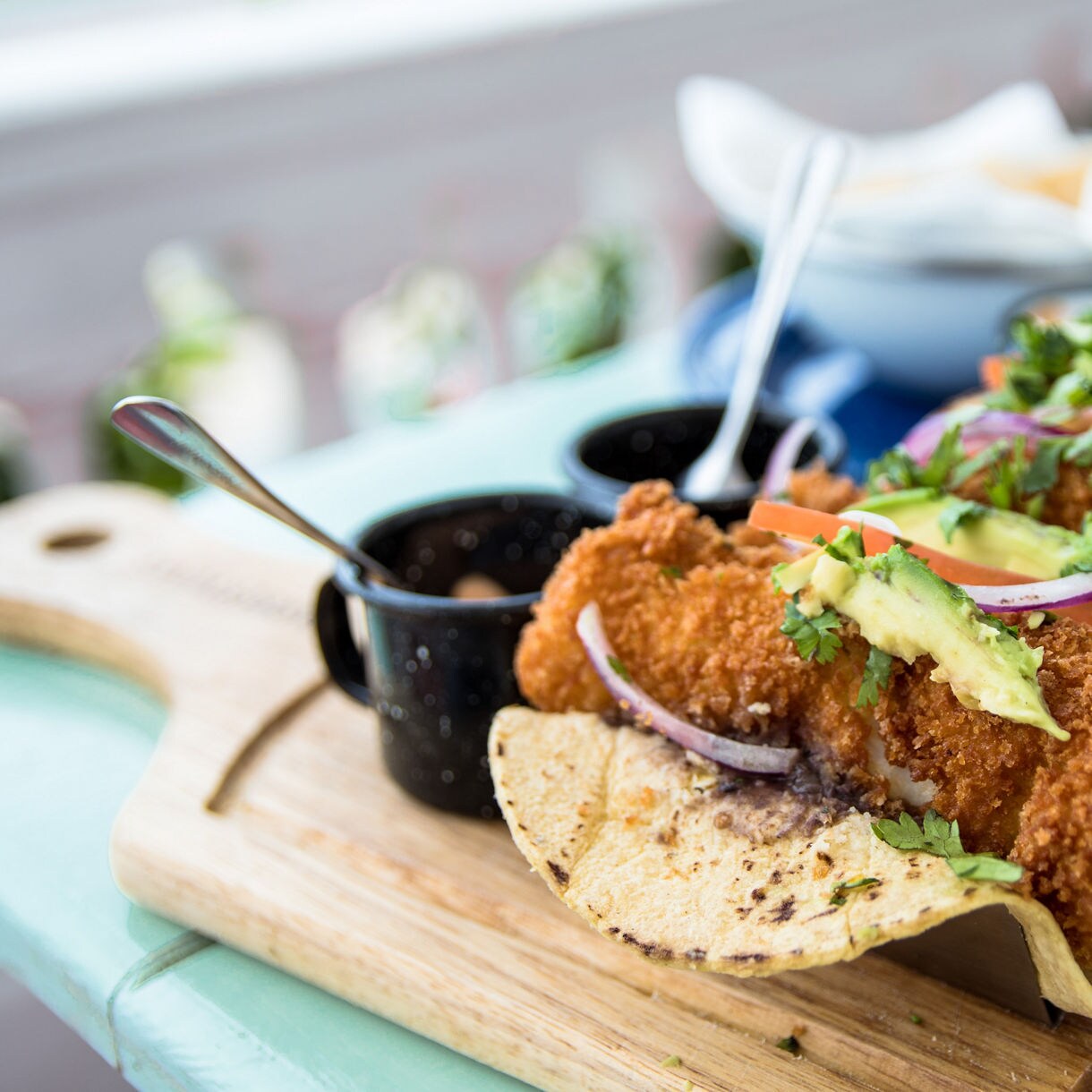 Three golden-brown breaded fish tacos served on a wooden board with slices of avocado, red onion and fresh cilantro on a light blue table.