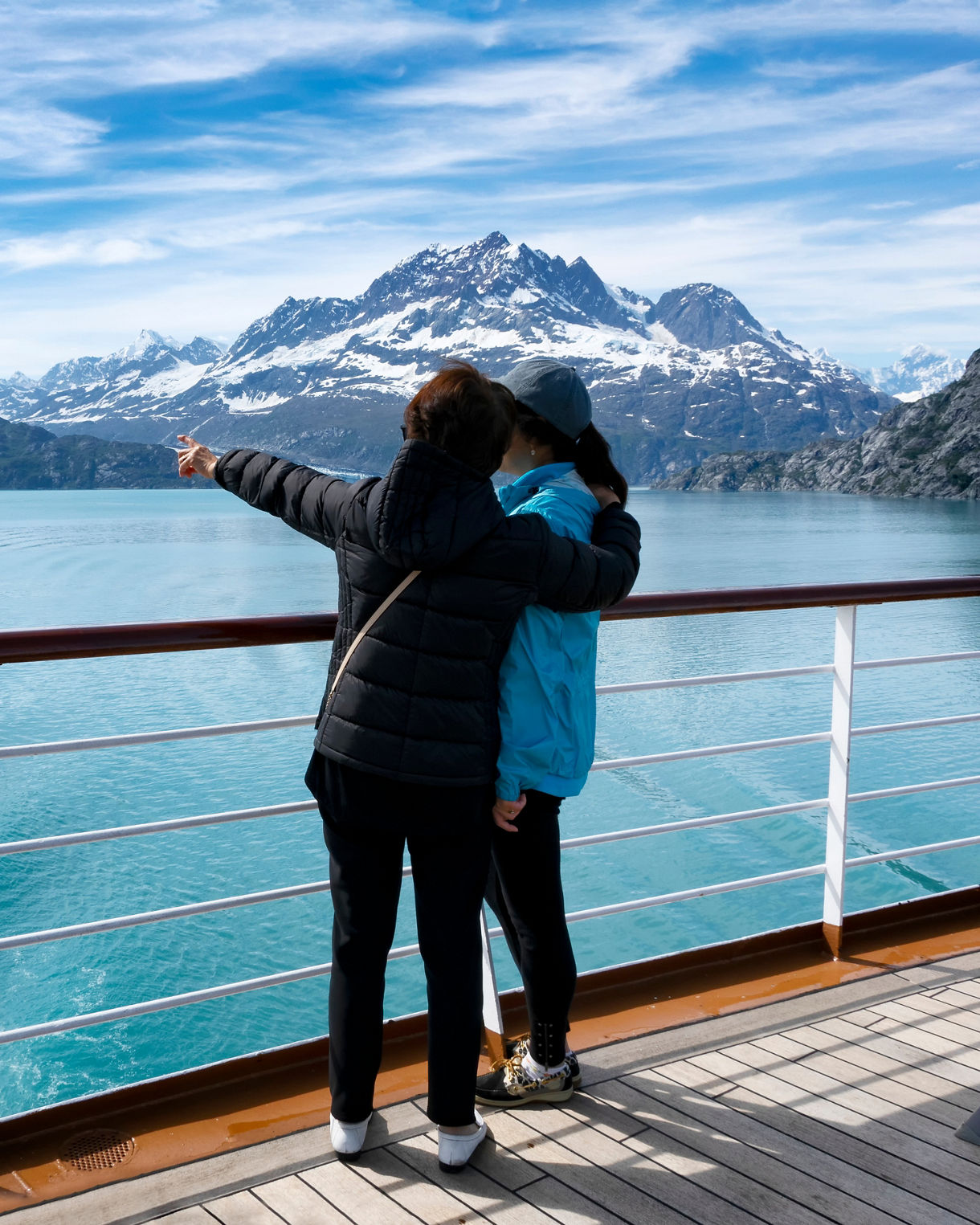 Two people stand on the deck of a ship, embracing as they look out over a calm turquoise fjord. Snow-capped mountains rise dramatically in the distance under a partly cloudy blue sky. The ship’s railing and wooden deck frame the foreground, emphasizing the vantage point from the cruise. The scene conveys a peaceful, contemplative moment in a cold, remote landscape.