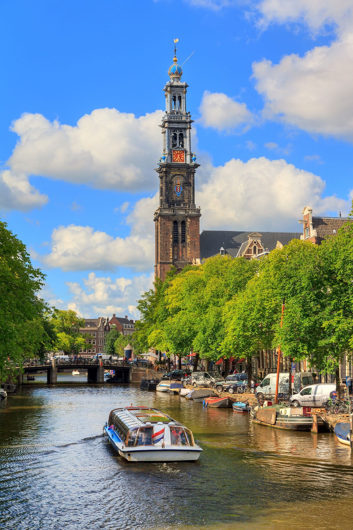 Sightseeing boat cruising down the Prinsengracht canal in Amsterdam with tree-lined streets, moored boats and the tall Westerkerk tower standing prominently in the background under a bright blue sky.