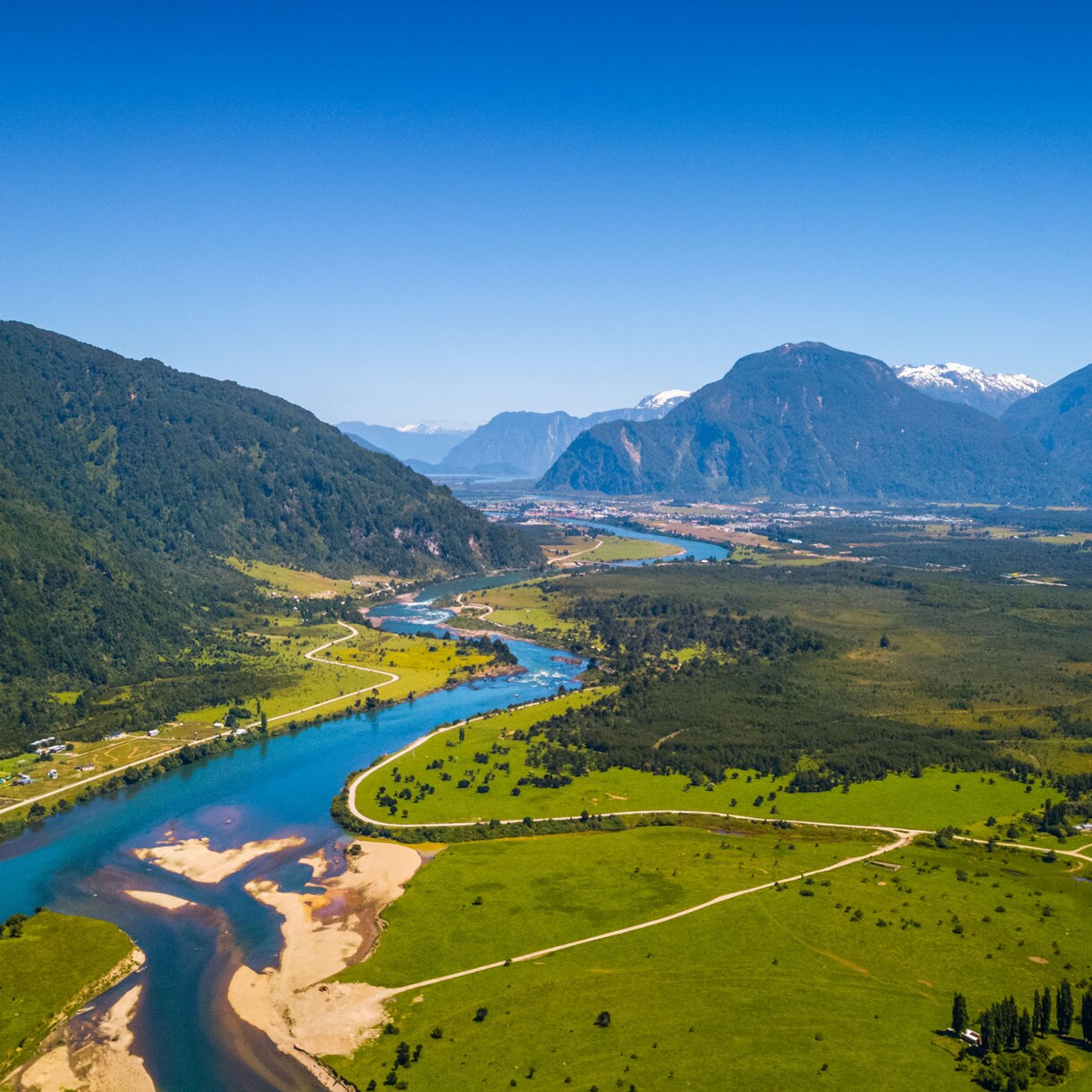 Aerial view of the Río Aysén flowing through bright green valleys with forested mountains and distant snow-capped peaks.