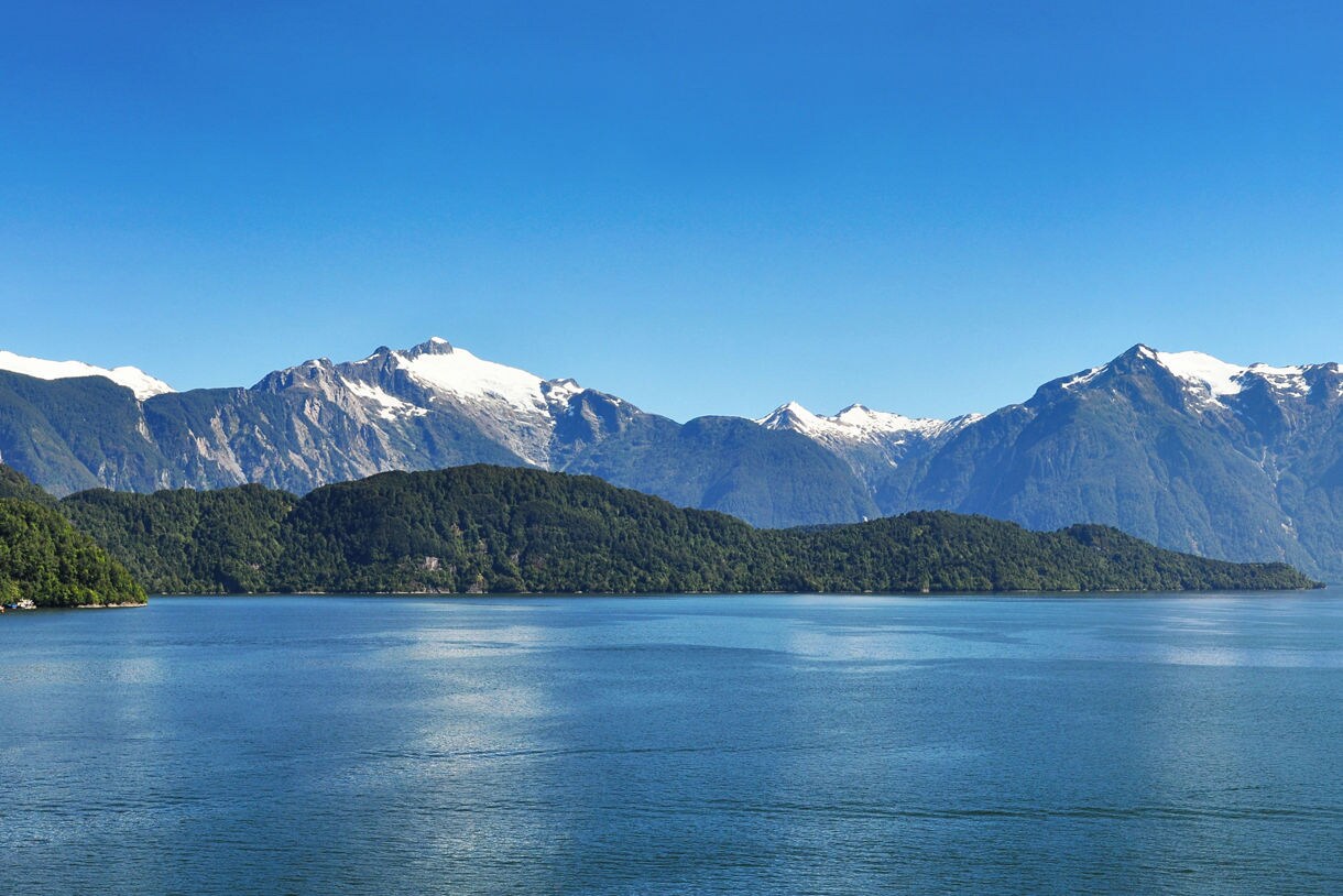 Wide view of Puerto Chacabuco’s calm blue water surrounded by forested hills and tall snow-capped mountains under a clear sky.
