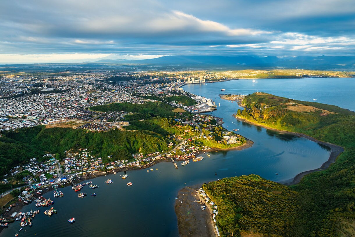 Aerial view of Puerto Montt showing a broad bay, clusters of fishing boats, forested hills and the city spreading across the coastline under soft evening light.