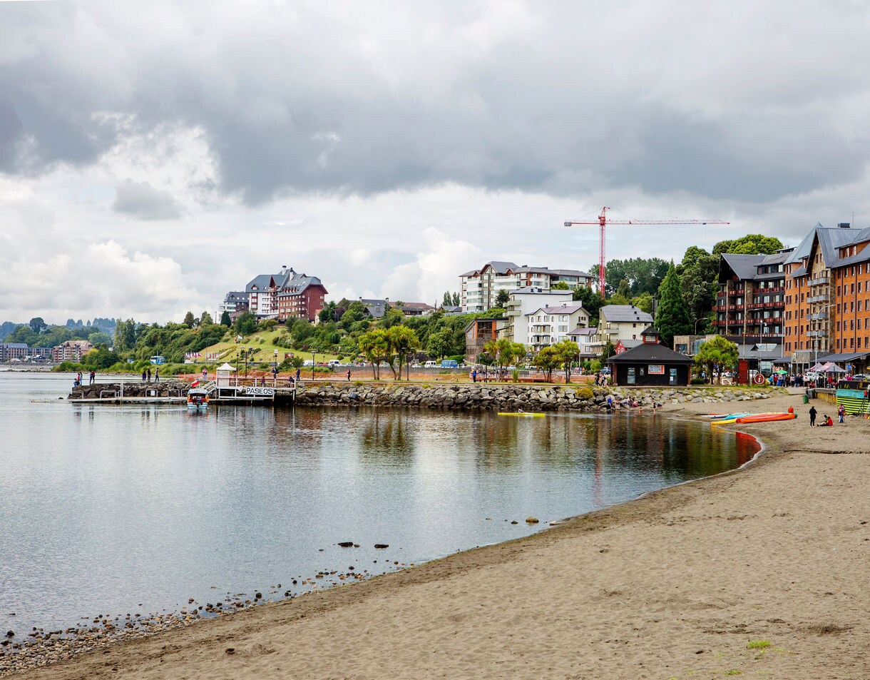 Sandy waterfront in Puerto Montt with calm water, small docks, colorful kayaks and hillside buildings beneath cloudy skies.