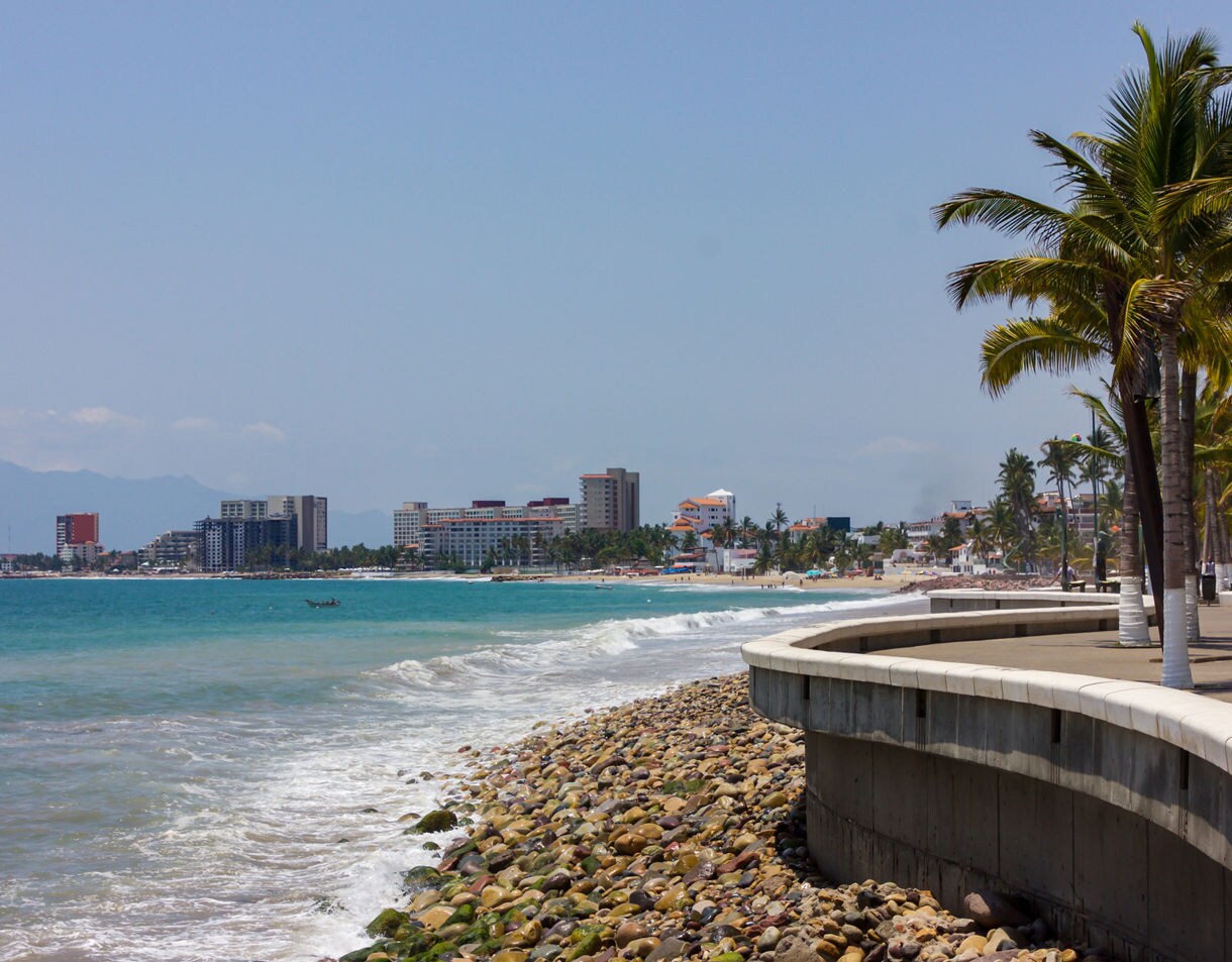 Palm-lined Puerto Vallarta Malecón with a curved seawall, rocky shoreline, turquoise waves and hotels rising along the coast.