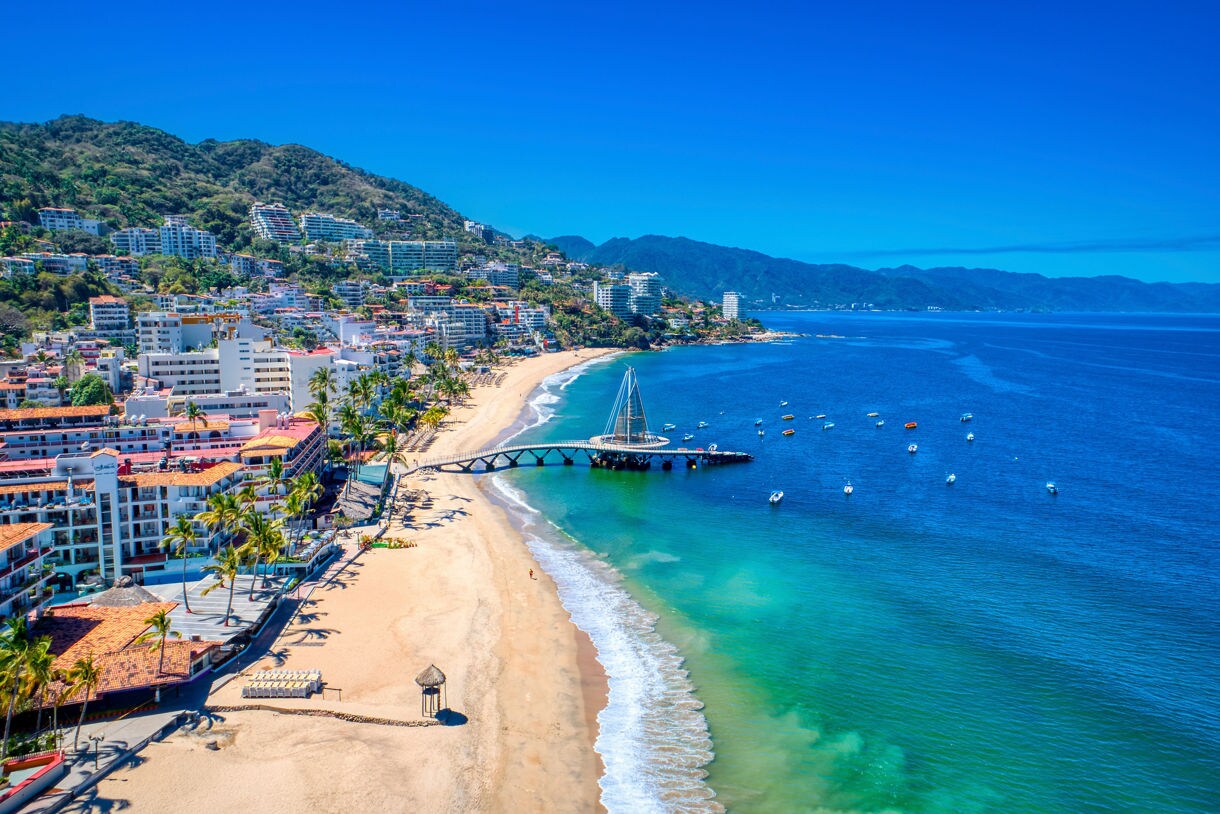Aerial view of Puerto Vallarta, Mexico, with sandy beach, modern pier extending into the ocean, boats on the water and lush green mountains in the background.