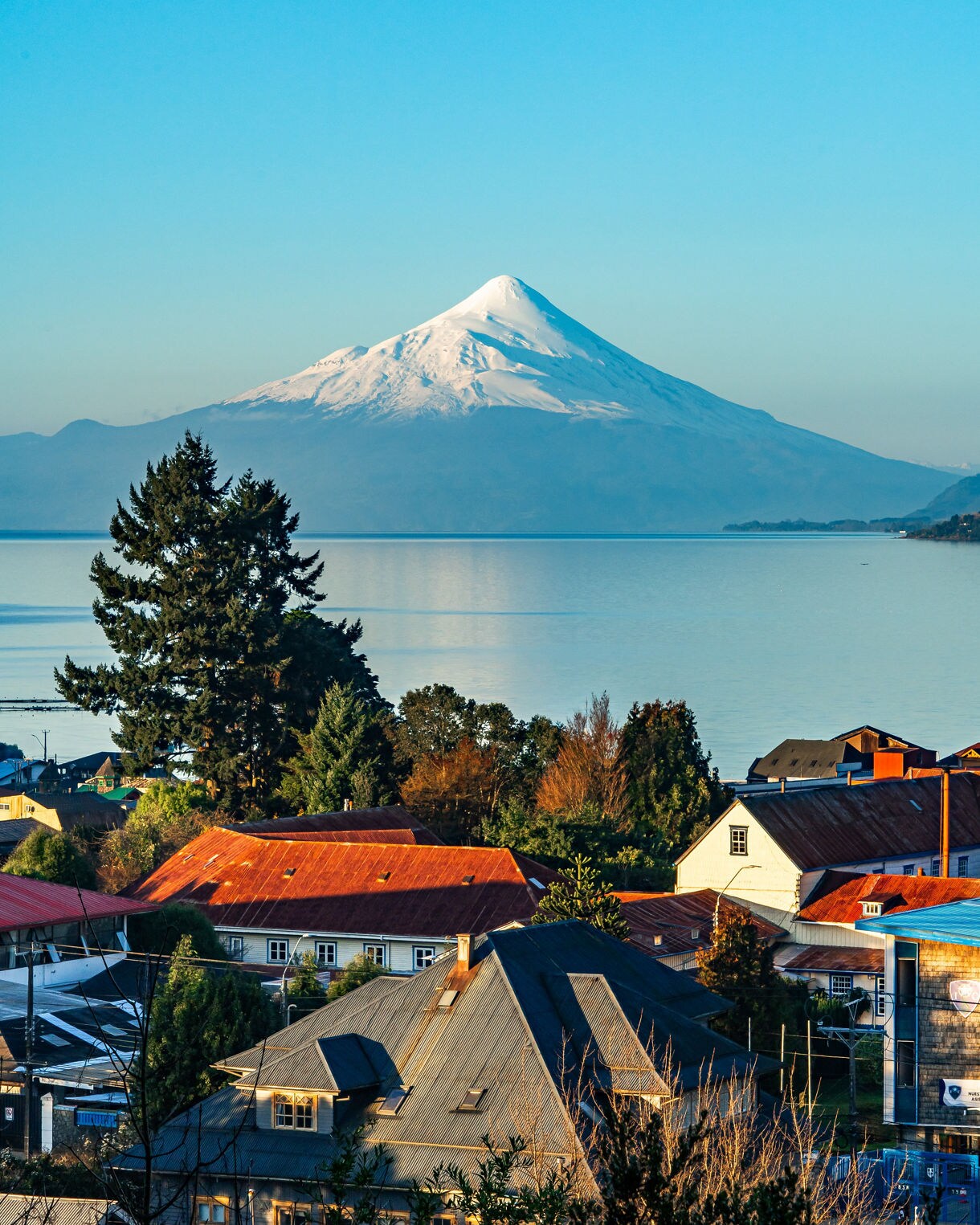 Snowcapped Osorno Volcano reflected in a still lake behind the rooftops and trees of Puerto Varas on a clear morning.