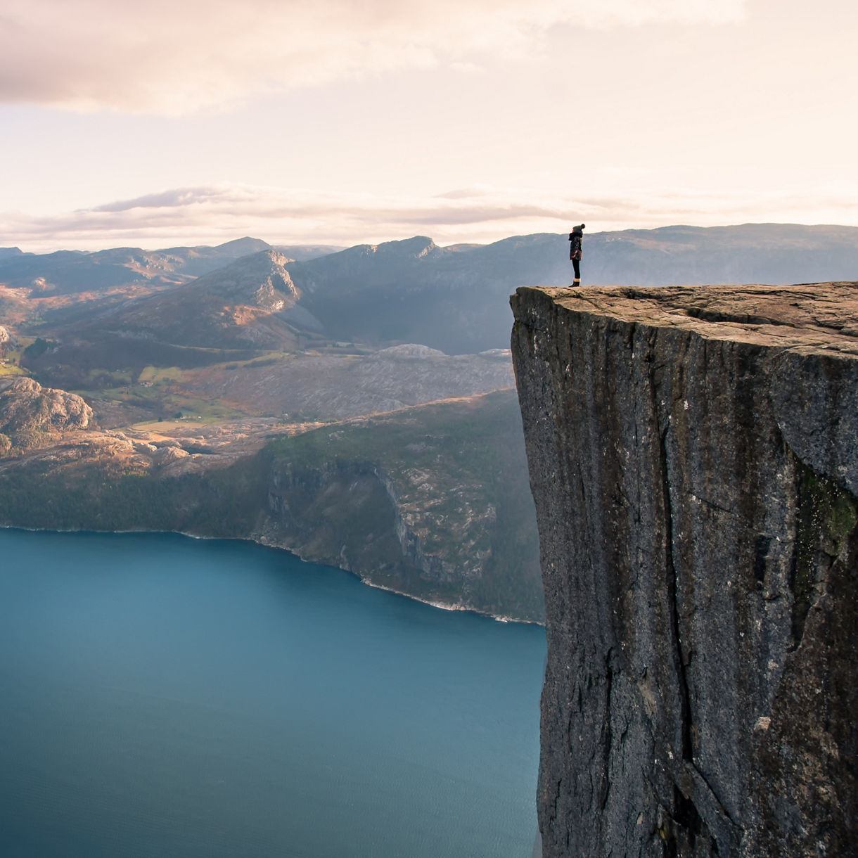 A lone hiker stands on the flat top of a sheer cliff high above a deep blue fjord with sunlit mountains stretching into the distance.