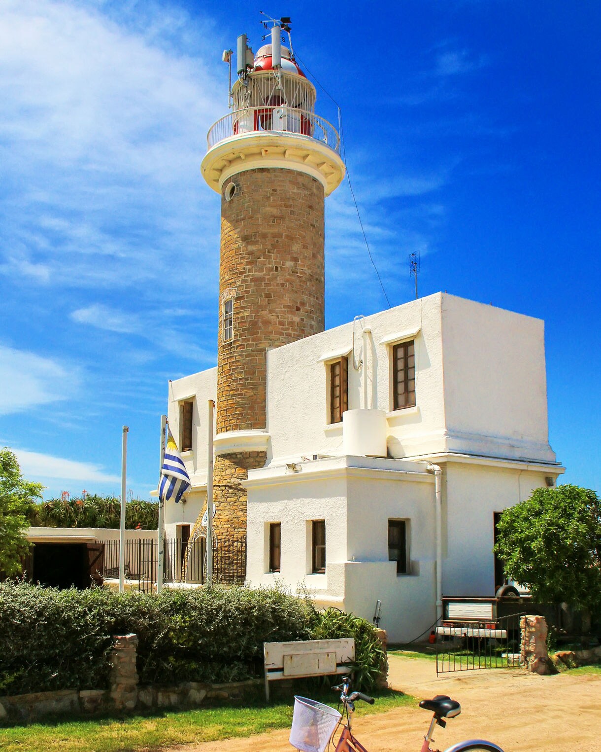 Stone lighthouse with a white keeper’s house beneath a bright blue sky, with a pink bicycle parked on a dirt path in the foreground.