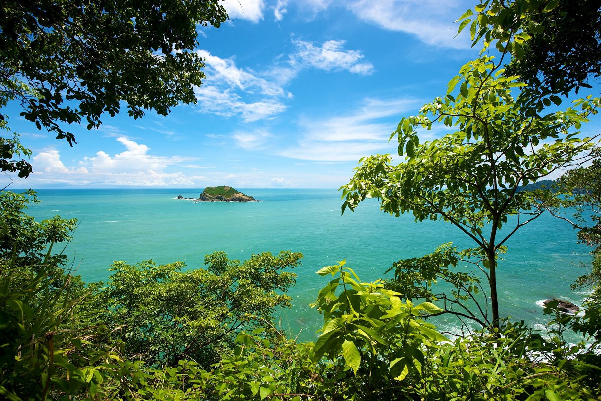 View of a small green island off the coast of Puntarenas, Costa Rica, framed by vibrant tropical foliage against a bright blue sky and turquoise ocean.