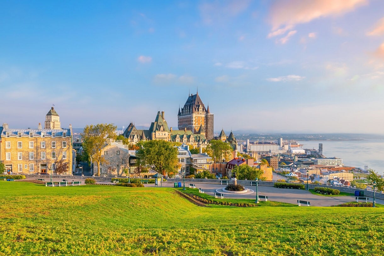 Elevated view of Old Québec with historic stone buildings, the Château Frontenac, lush green lawns and the Saint Lawrence River glowing under a soft sunset sky.