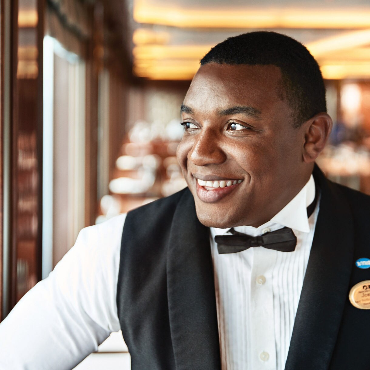 A male waiter is seen wearing a formal black vest, white shirt, and bow tie, standing in a well-lit restaurant hallway. The setting features polished wood panels and a softly blurred dining area in the background. The waiter has a visible name badge and a blue pin on his vest. The overall mood is professional and welcoming, with warm lighting enhancing the elegant atmosphere.