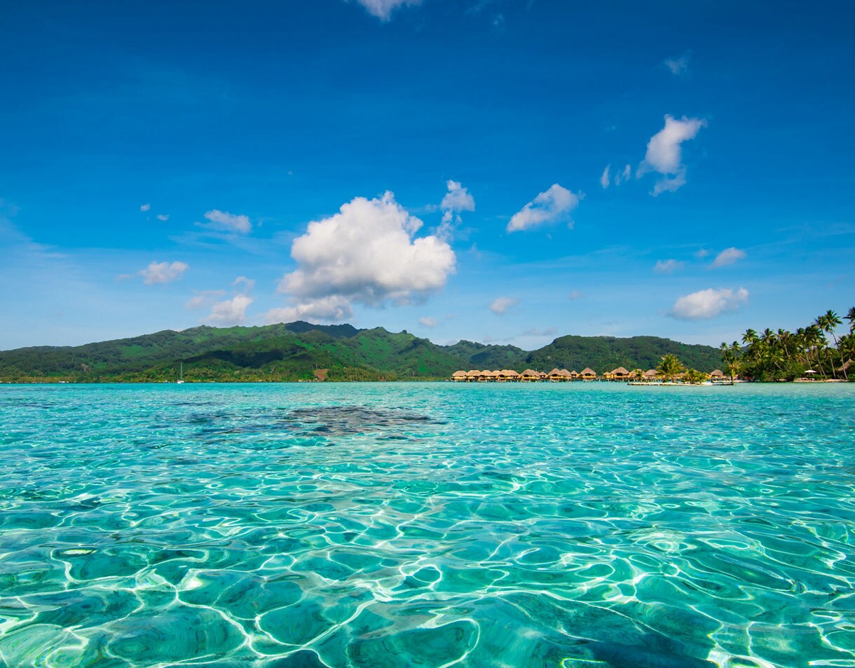 Shallow turquoise lagoon with crystal reflections, backed by green mountains and palm-fringed shores on Raiatea.