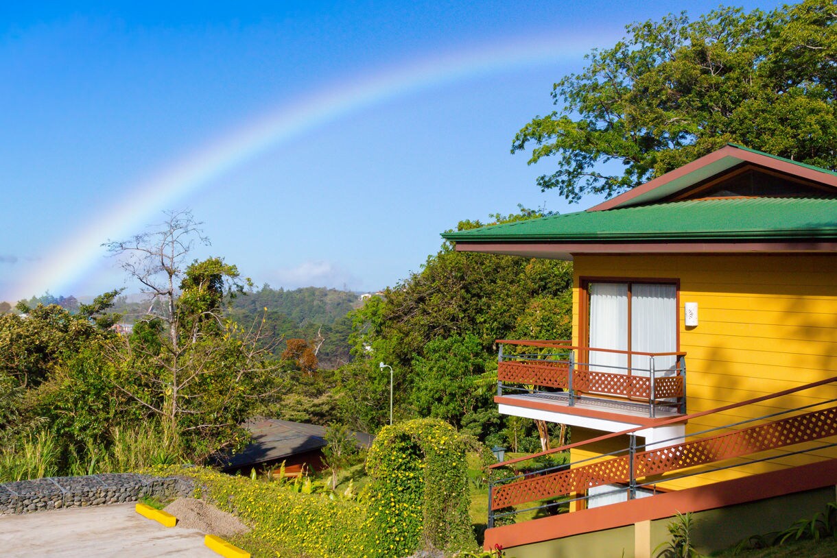 Yellow bungalow with green roof and balcony set against a forested hillside and a rainbow stretching across the blue sky.