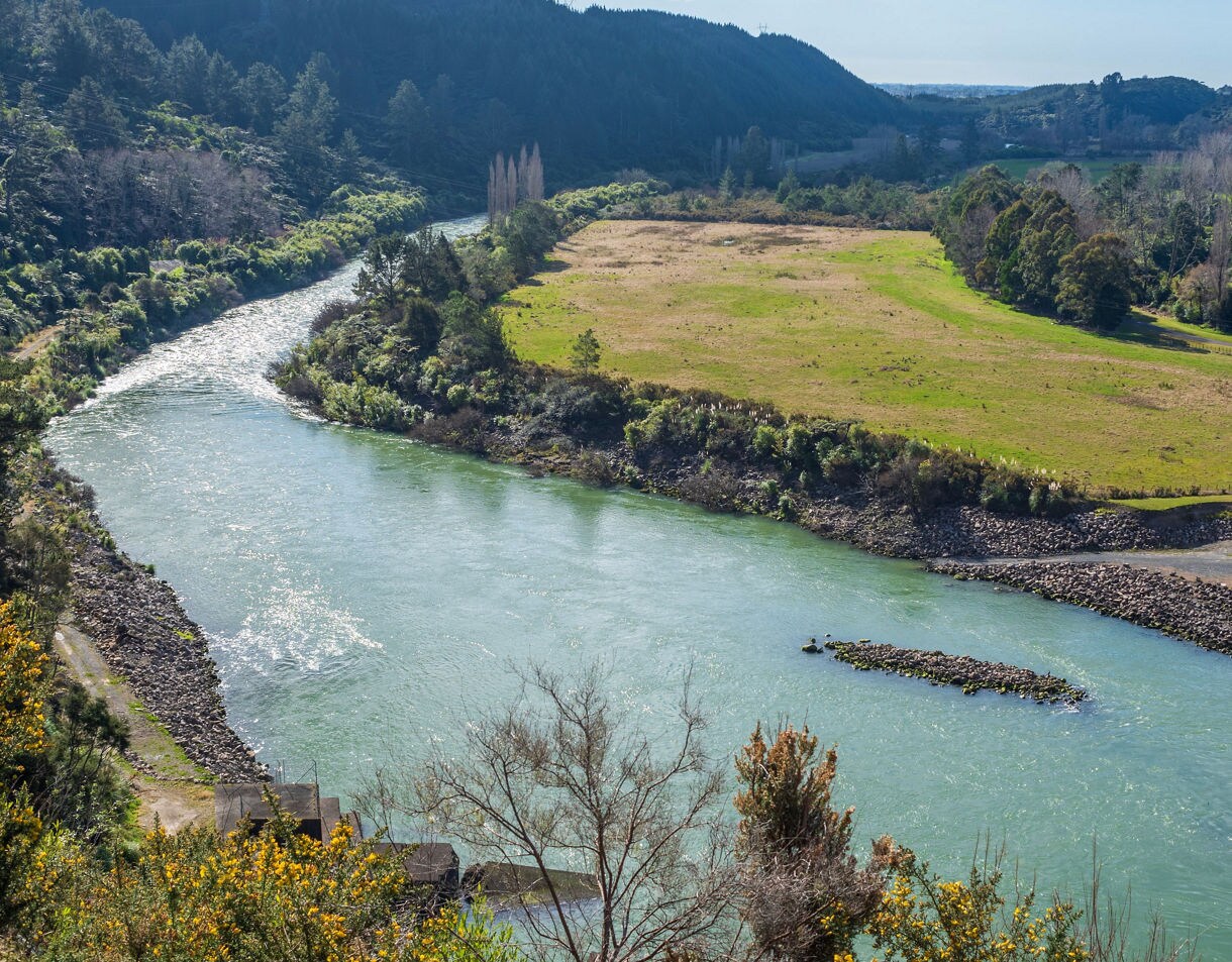 A wide, gently curving river bordered by grassy fields and forested hills under clear daylight.