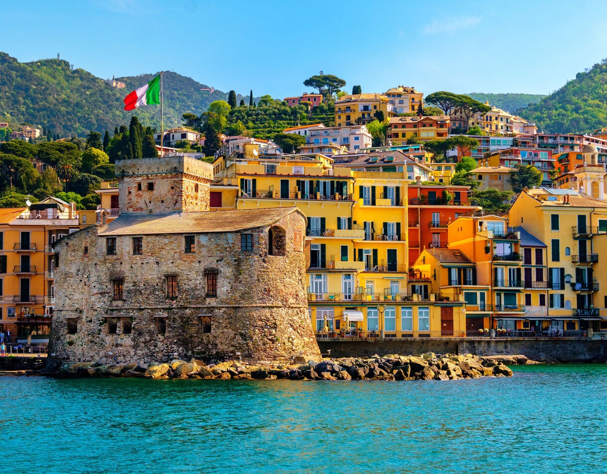 Stone castle flying the Italian flag on Rapallo’s waterfront, surrounded by colorful hillside buildings and turquoise water.