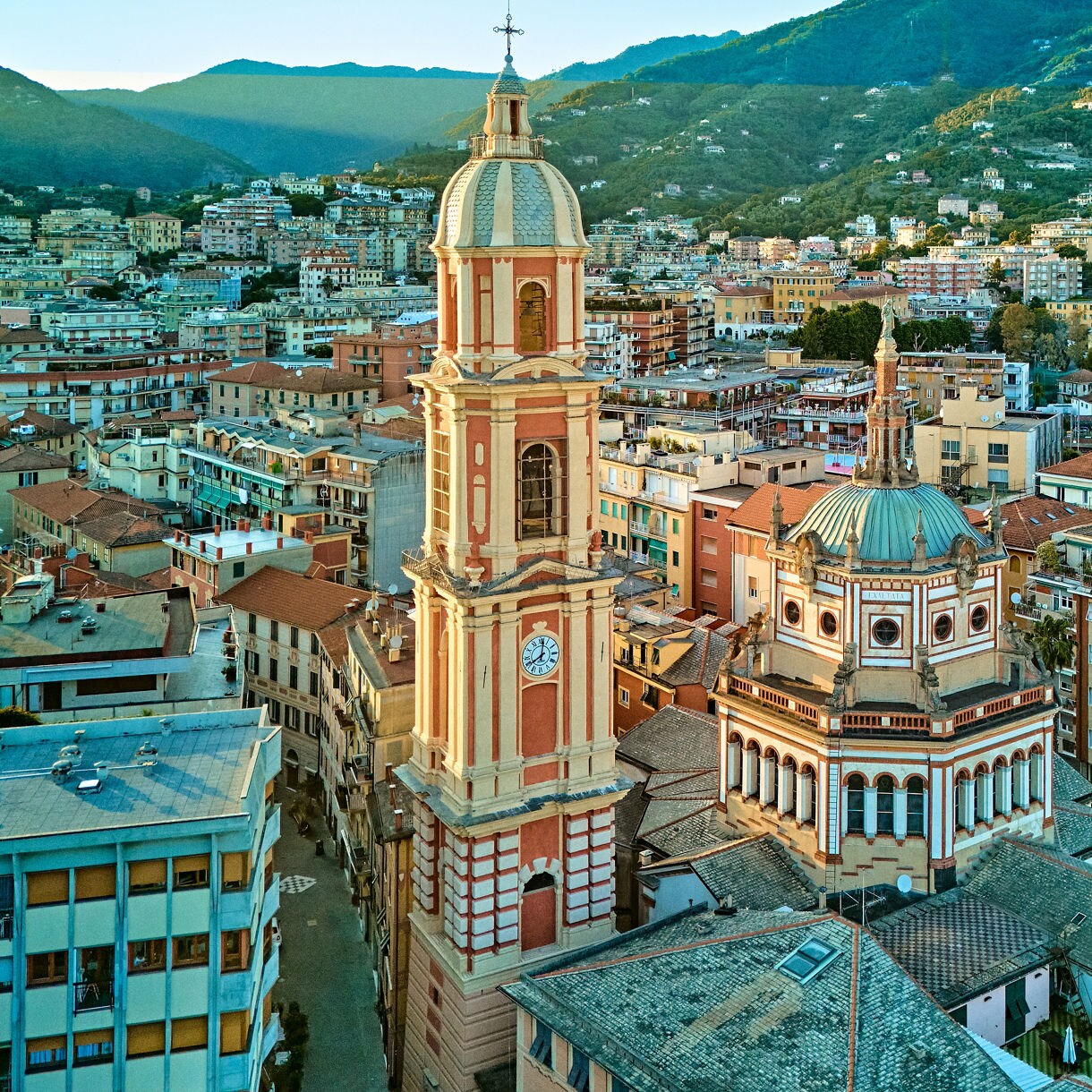 Aerial view of Rapallo showing a tall bell tower and domed church surrounded by pastel buildings, with green hills rising in the background.