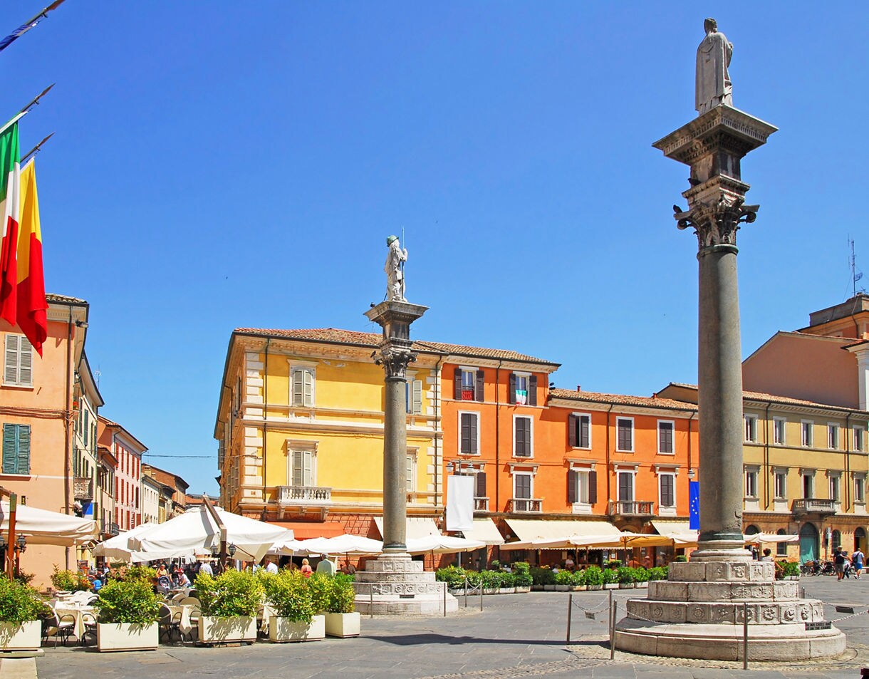 iazza del Popolo in Ravenna featuring colorful historic buildings, two stone columns topped with statues, outdoor café seating and Italian and European Union flags beneath a clear blue sky.