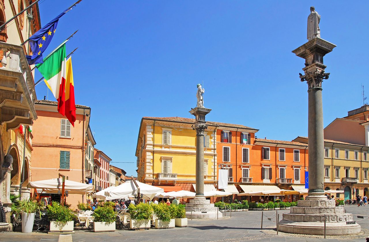 Piazza del Popolo in Ravenna featuring colorful historic buildings, two stone columns topped with statues, outdoor café seating and Italian and European Union flags beneath a clear blue sky.
