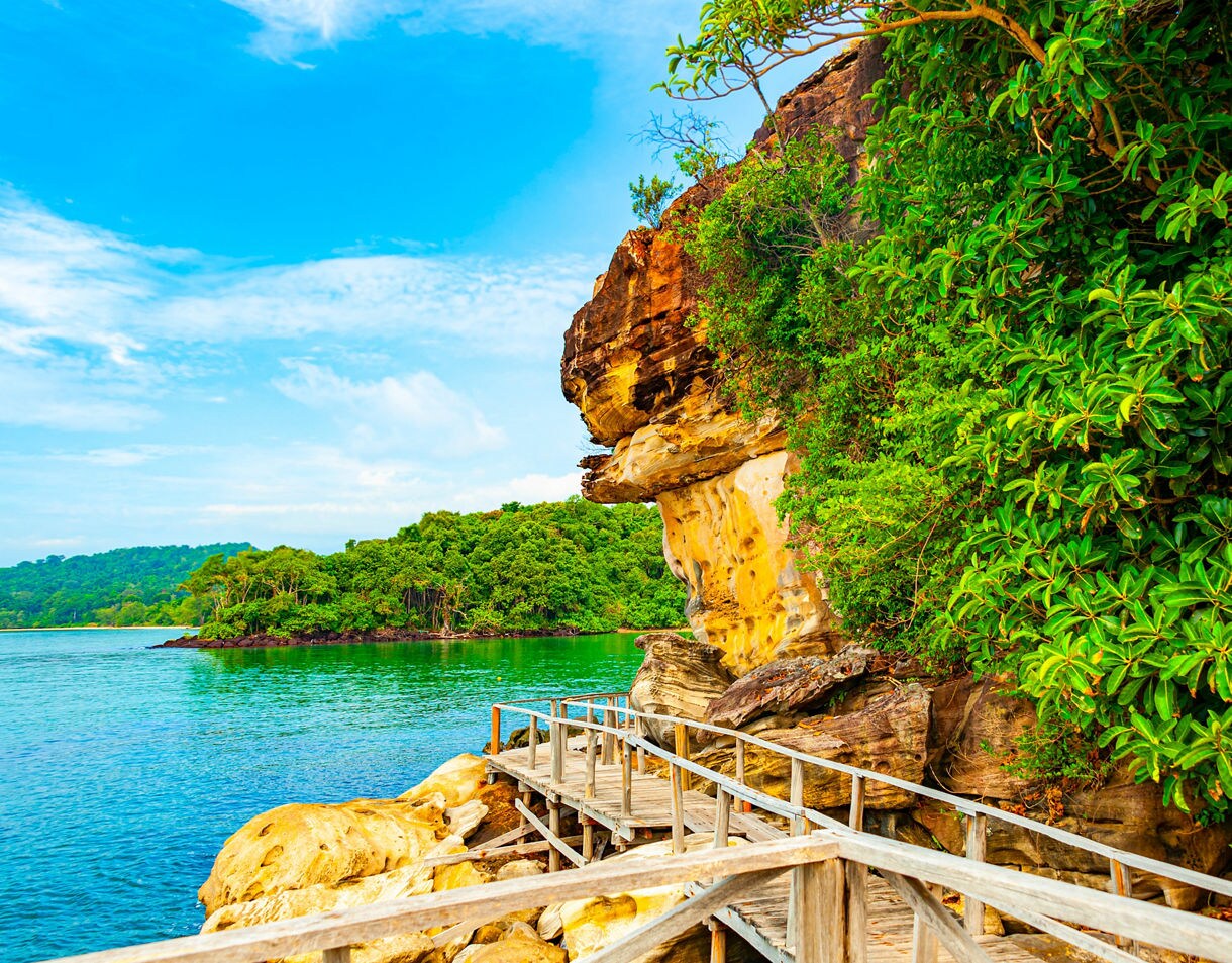 A wooden boardwalk curves along colorful sandstone cliffs and dense green foliage above clear turquoise water.