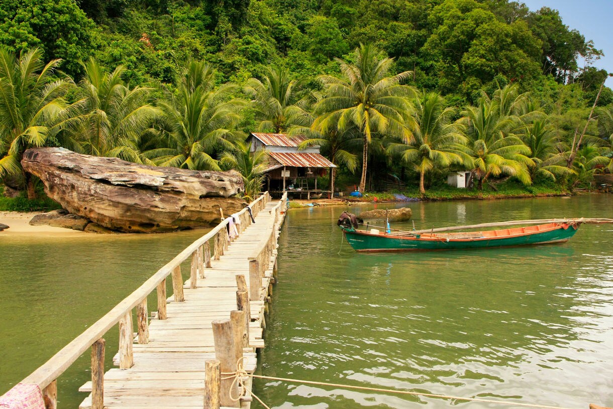 Wooden walkway leads to a small hut beside calm green water with palm trees, a large rock formation and a moored wooden boat.