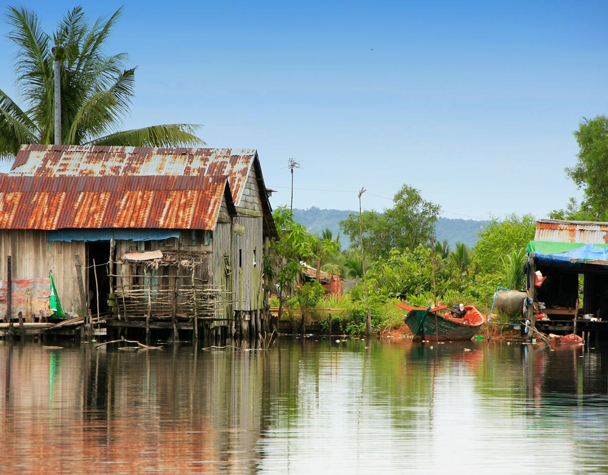 Rustic wooden stilt house with a rusted tin roof stands beside calm reflective water with small boats and lush greenery nearby.