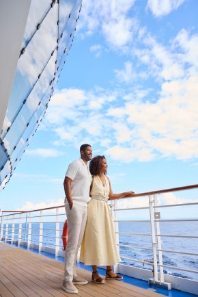 A couple stands together on the deck of a cruise ship, smiling as they enjoy the ocean view under a bright blue sky with scattered clouds.