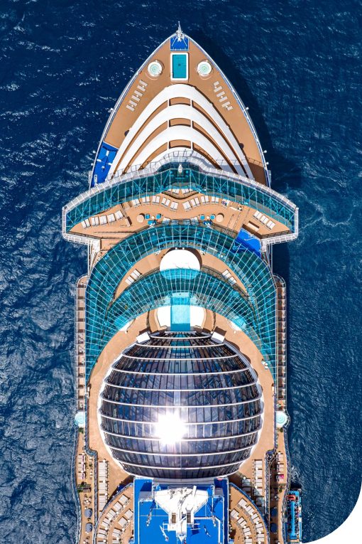 Aerial view of a large cruise ship's top deck, featuring multiple pool areas, lounge chairs, and glass-domed structures, sailing through deep blue ocean waters.