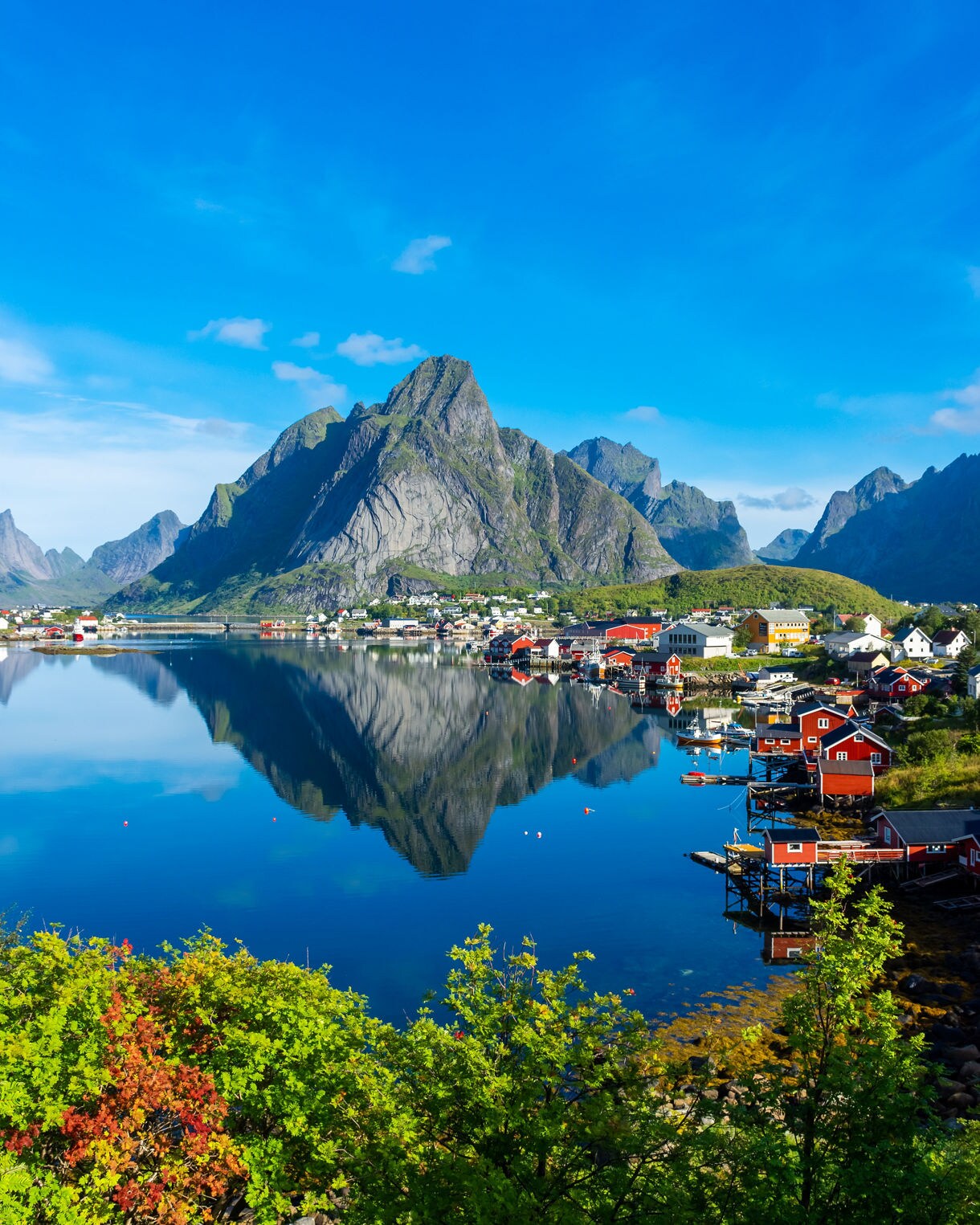 Red fishing cabins and houses along the water’s edge in Reine, Norway, with steep rocky mountains reflected in the calm fjord.